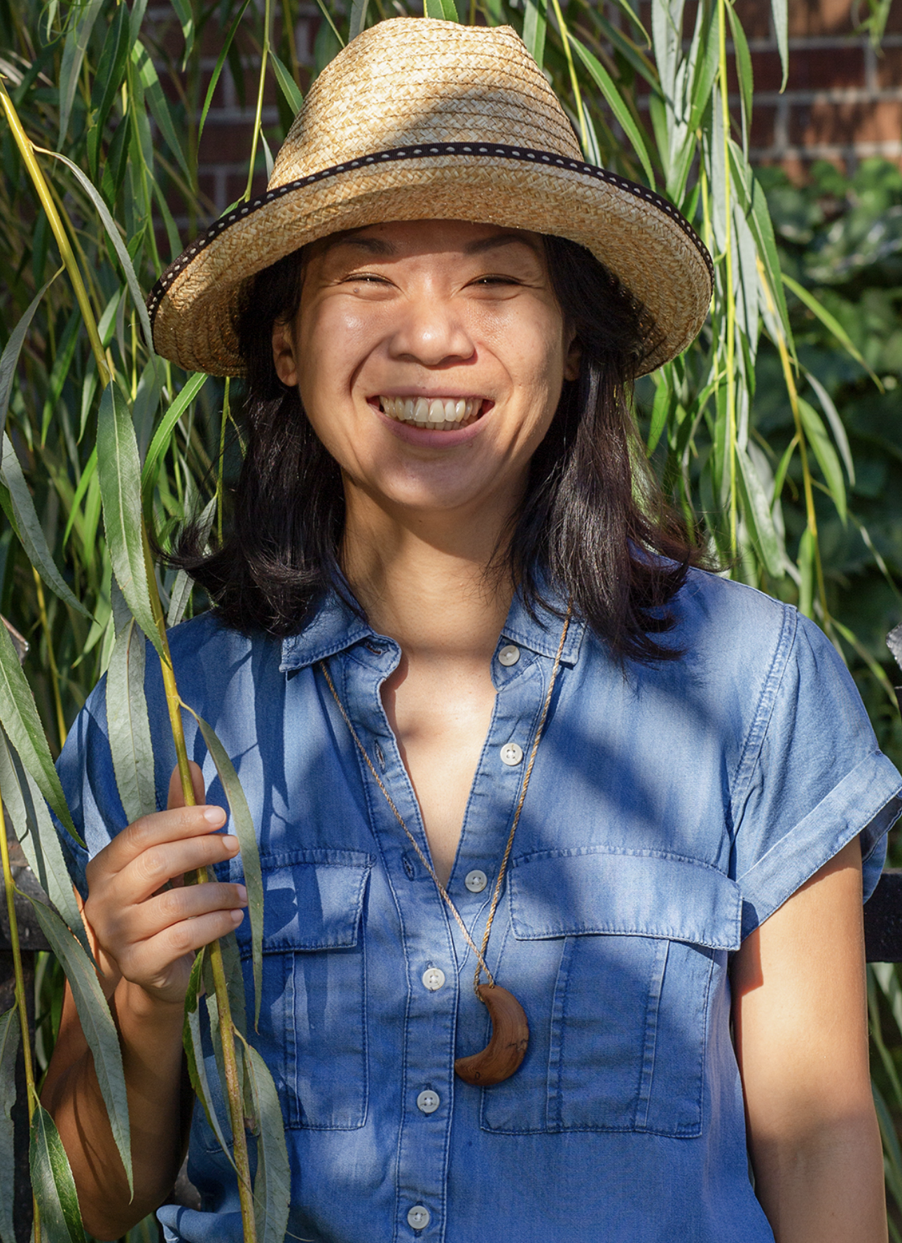 South Asian person wearing a straw hat, blue short sleeve button-up smiling at the camera.